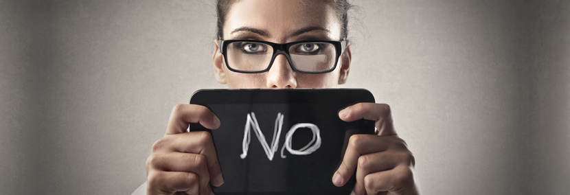 Image of business woman holding a sign saying no. She's wearing black reading glasses peering over the sign.