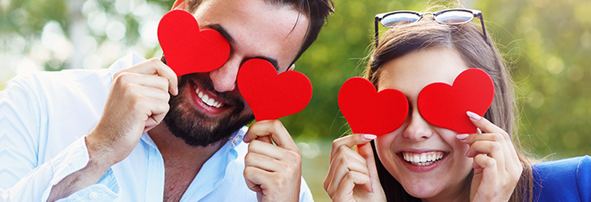 man with dark hair holding up valentines day hearts to eyes and a woman with long hair holding valentines day hearts to eyes