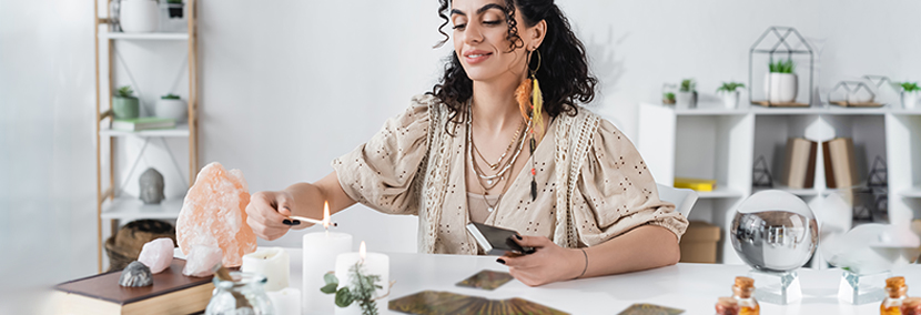 a psychic with black curly hair sitting in front of her reading desk lighting a candle. A tarot card spread and candles appear on the table