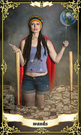 Two of Wands tarot card featuring a woman in casual clothing and a red cape, holding a smoking stick in one hand and a globe balloon in the other, standing between two wands amid stone ruins.